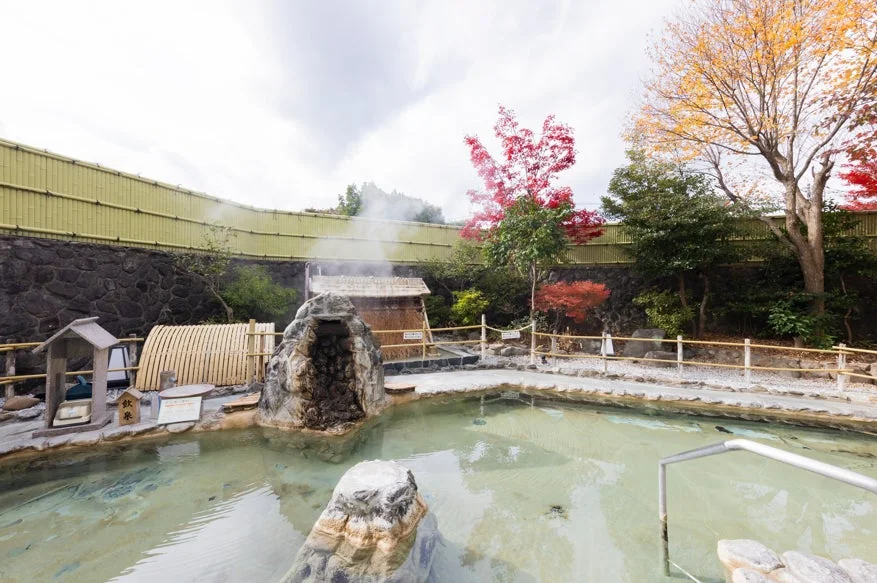 湯気が立ち上る露天風呂の風景です。周囲には赤や黄色に色づいた木々があり、秋の風情を感じさせます。竹垣に囲まれ、落ち着いた雰囲気の日本らしい温泉です。