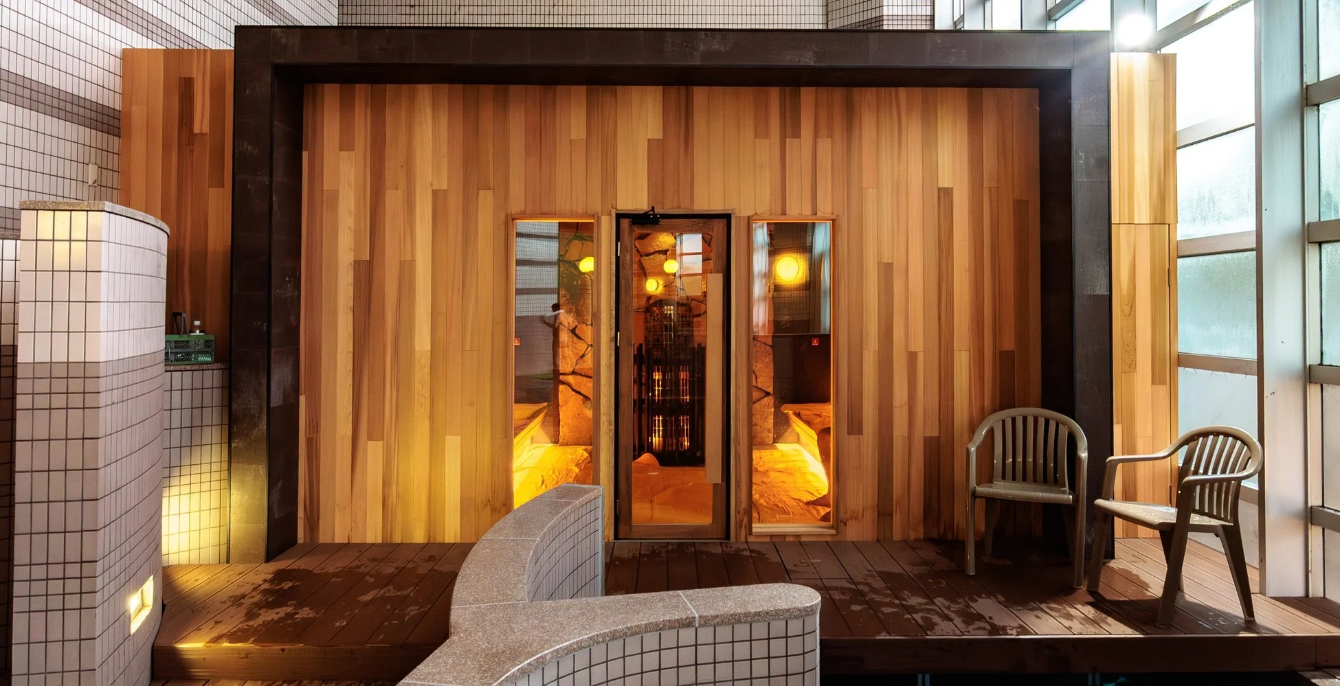 モダンな温浴施設の一角で、木目調の壁に囲まれたサウナルームが中央に位置しています。濡れた木製デッキとタイル張りのベンチがあり、リラックスできる空間が広がっています。