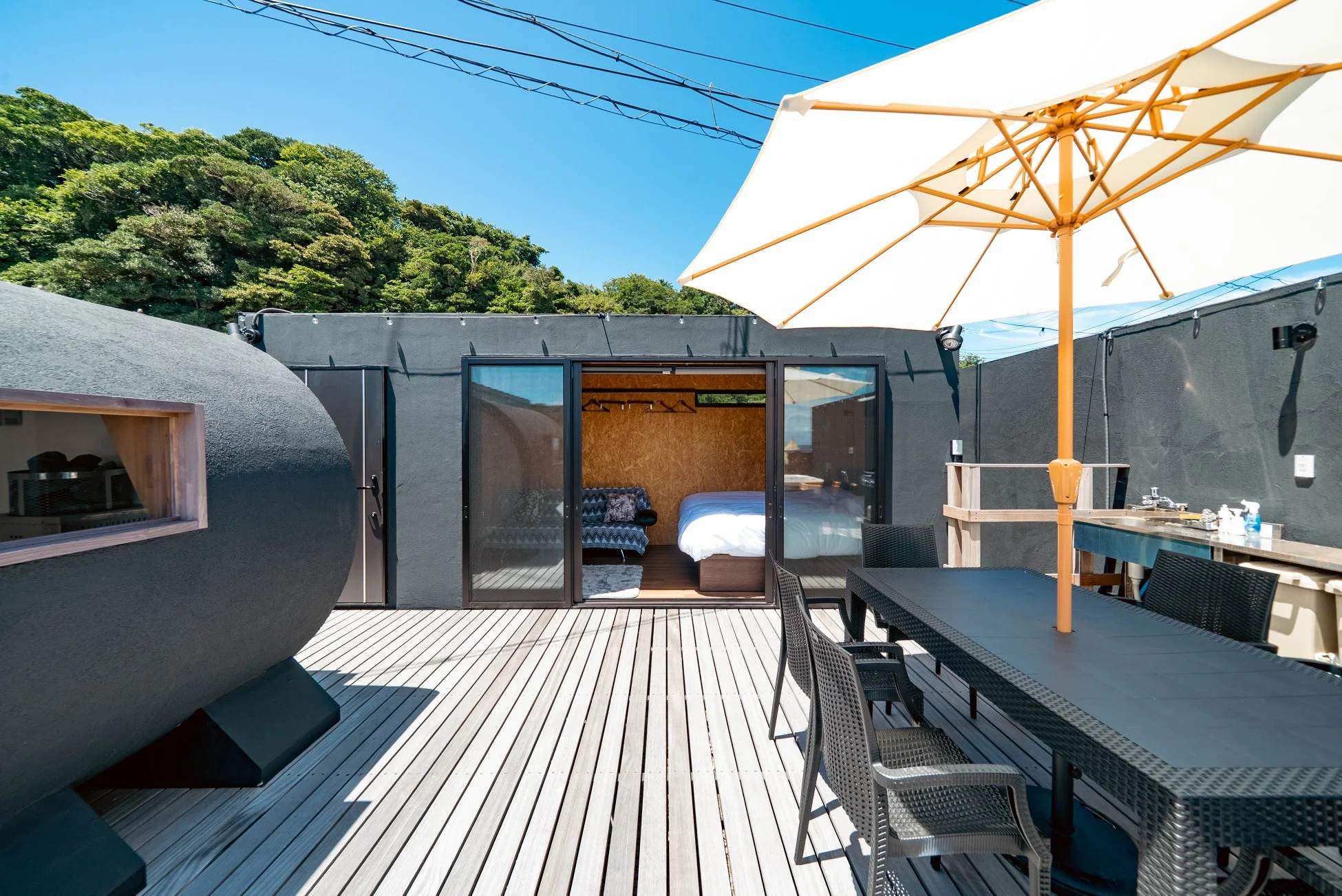 青空の下、広々としたウッドデッキにモダンな宿泊施設、屋外ダイニング、白いパラソルが配置されたリラックスできる空間