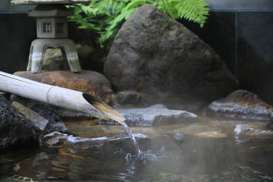 竹の筒から温かい水が流れ落ち、湯気が立ち込める和風の温泉風景