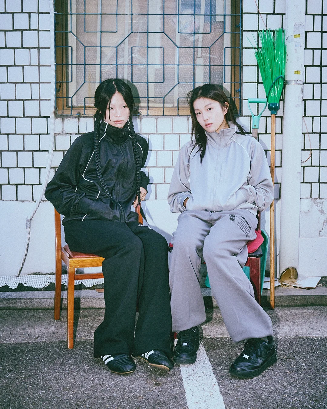 タイル張りの壁と窓を背景に、トラックスーツを着た若い女性が二人椅子に座っています