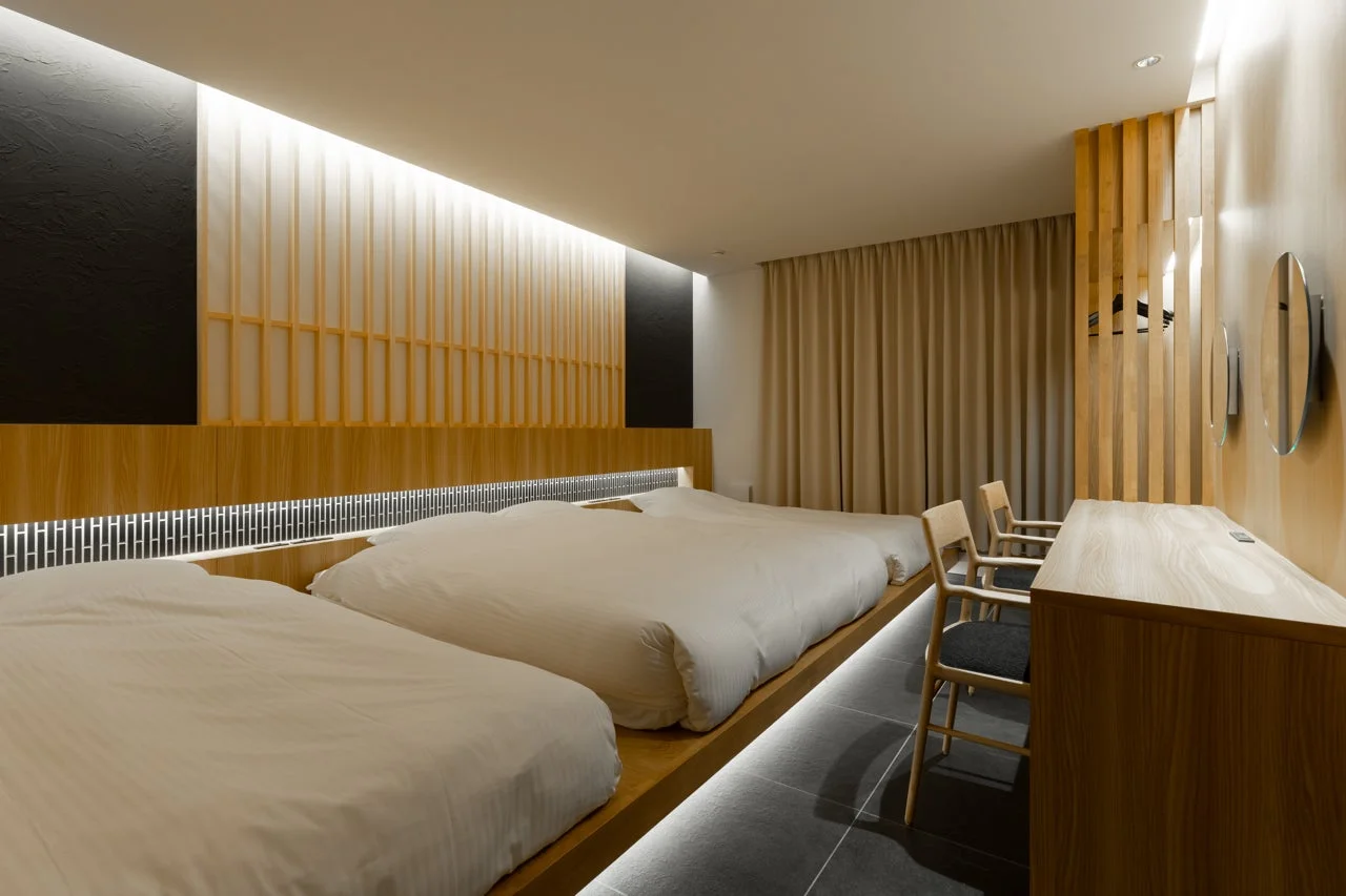 和モダンなデザインの寝室で、木製の低いベッドが2台置かれています。間接照明が落ち着いた雰囲気を演出し、木製家具と障子風の壁が特徴的です。