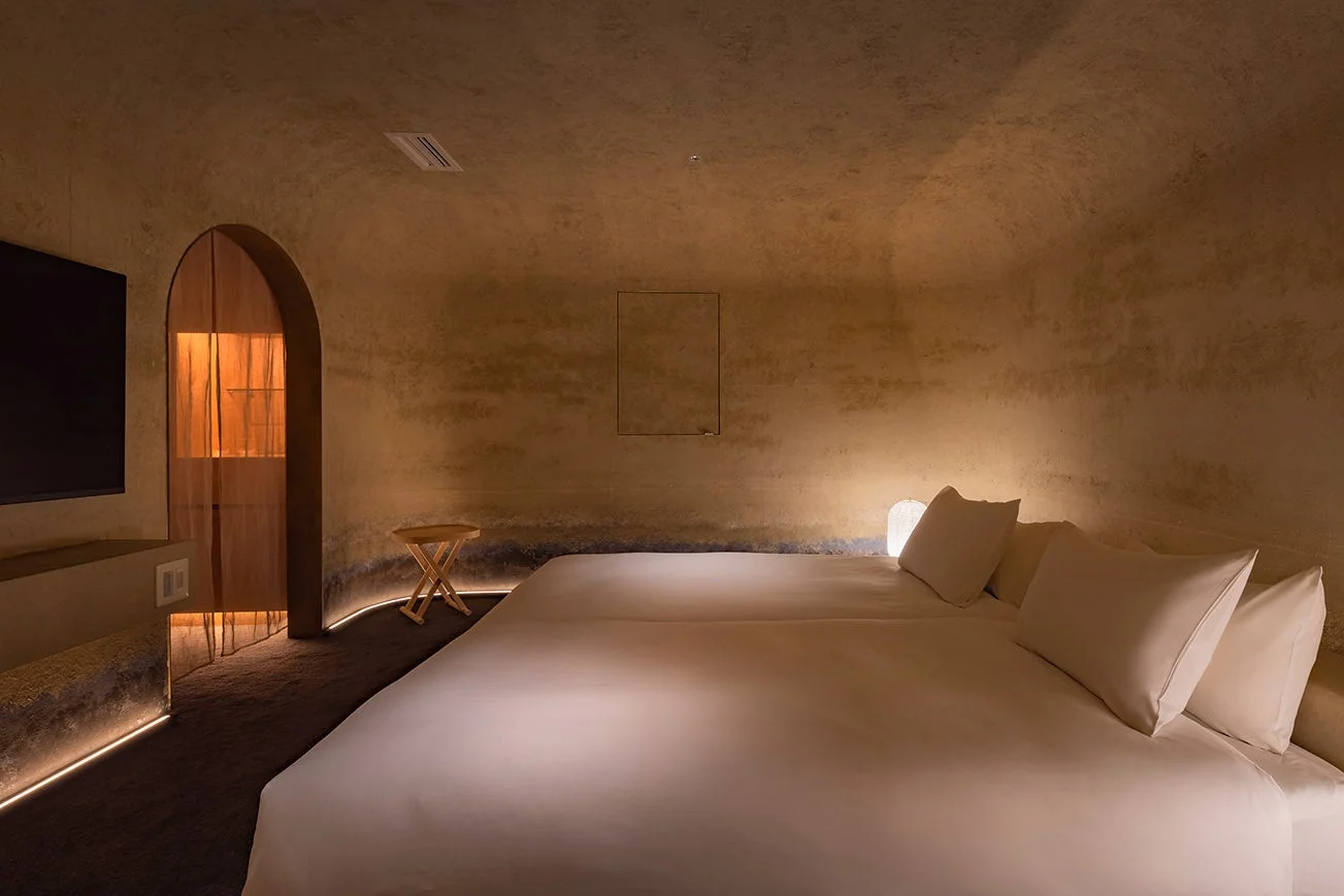 温かみのある照明が特徴的な、土壁のような質感の壁に囲まれたモダンな寝室です。ツインベッドが配置され、足元には間接照明があり、奥にはアーチ型の入り口が見えます。