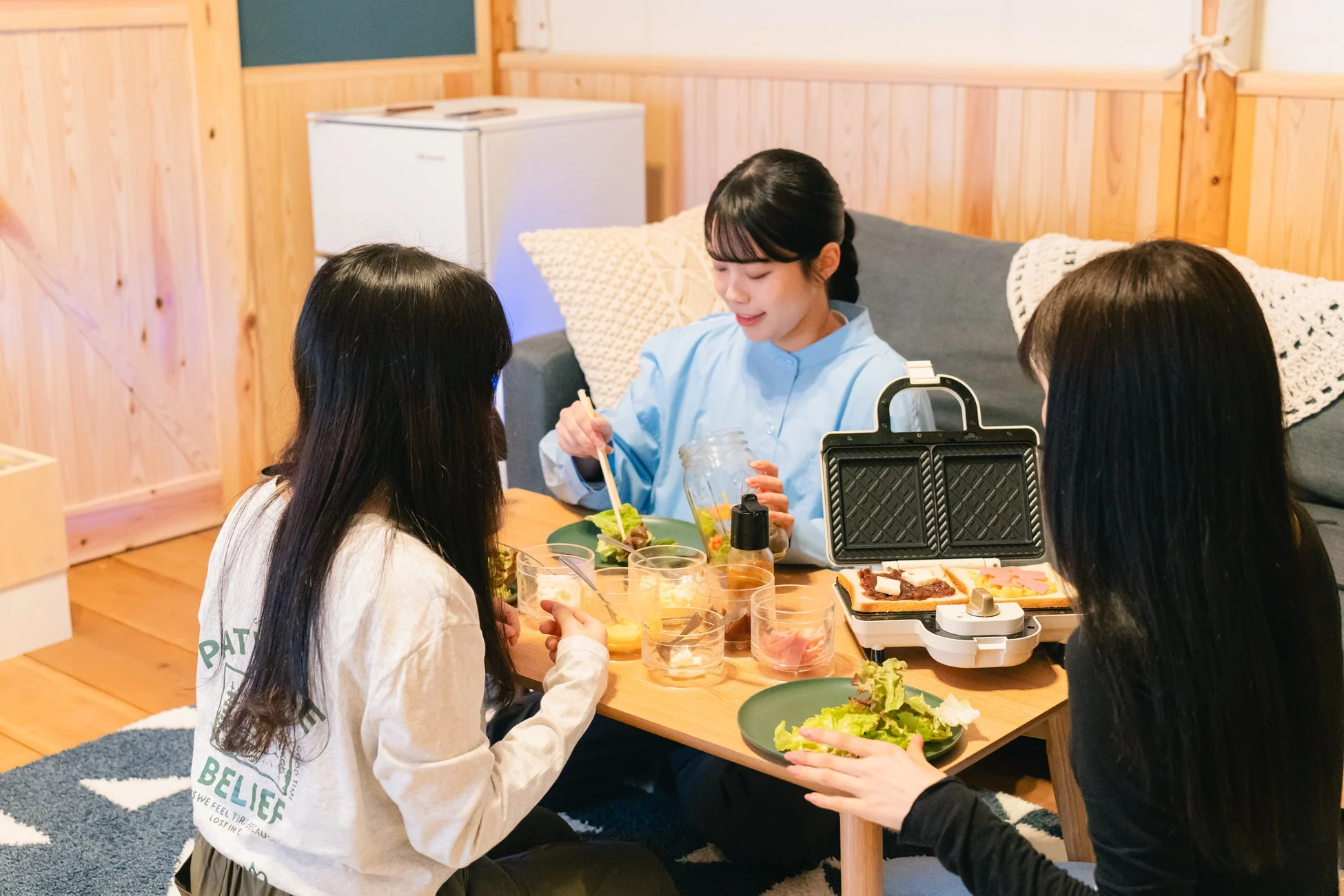 リビングで3人の女性がホットサンドメーカーを使ってホットサンドを作りながら、サラダと共に食事を楽しんでいる様子