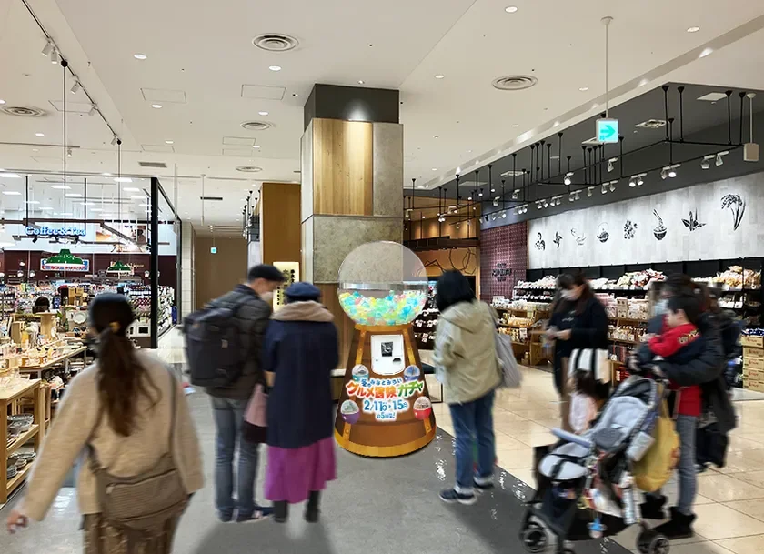 商業施設内の食料品売り場で、多くの買い物客が行き交う中、大きな「グルメ冒険ガチャ」が設置されている様子。