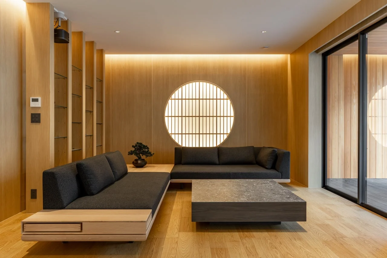 明るい木材と落ち着いたグレーを基調とした、和モダンなリビングルームです。円形の障子風照明が特徴的で、L字型に配置されたソファと大きなコーヒーテーブルが置かれています。