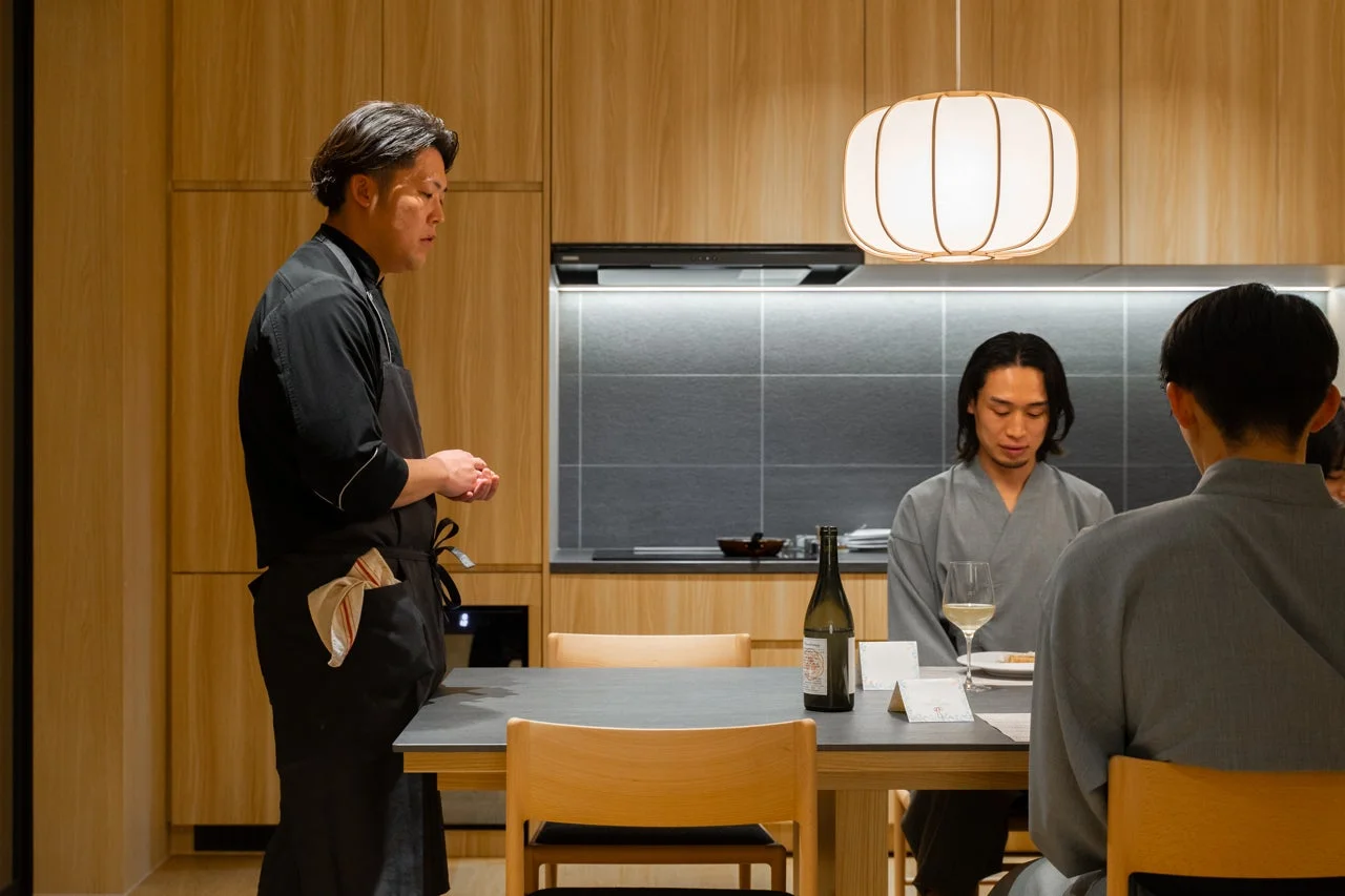 モダンなダイニングスペースで、シェフが二人のゲストに何か説明をしている様子。テーブルには日本酒のボトルとグラスが置かれ、落ち着いた雰囲気の中で食事が提供されている光景です。