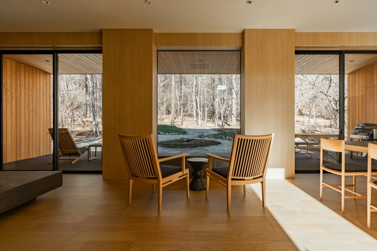 広々とした窓から冬の森を望むモダンな室内空間です。木製の家具とミニマリストなデザインが特徴で、中央の窓の外には枯山水のような庭が見えます。自然光が差し込む落ち着いた雰囲気です。