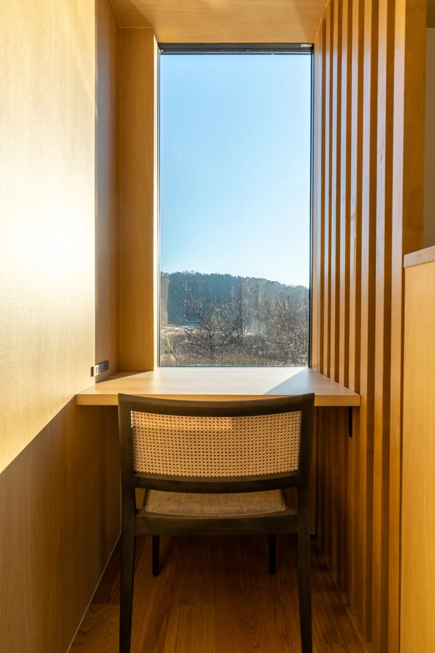 自然光が差し込む木製のモダンなワークスペース。大きな窓から青空と遠景が見え、シンプルなデスクとラタン調の椅子が配置された落ち着いた空間。