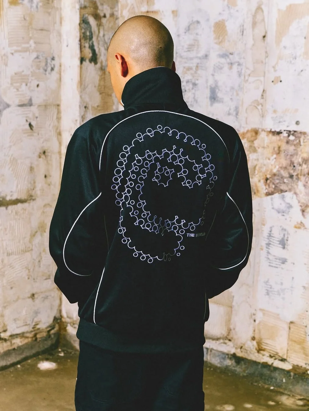 坊主頭の男性が、背中に円形の幾何学模様と「THE WORLD」のロゴがデザインされた黒いトラックジャケットを着て立っている後ろ姿