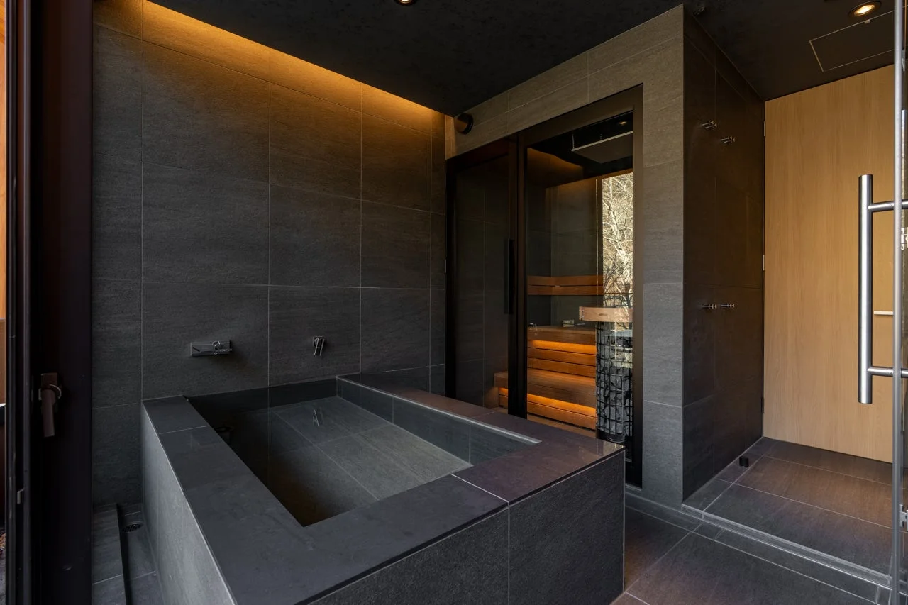 黒を基調としたモダンで高級感のある浴室。石造りのバスタブと、ガラス越しに自然の景色が広がるプライベートサウナが特徴的です。温かい間接照明が落ち着いた贅沢なリラックス空間を演出しています。