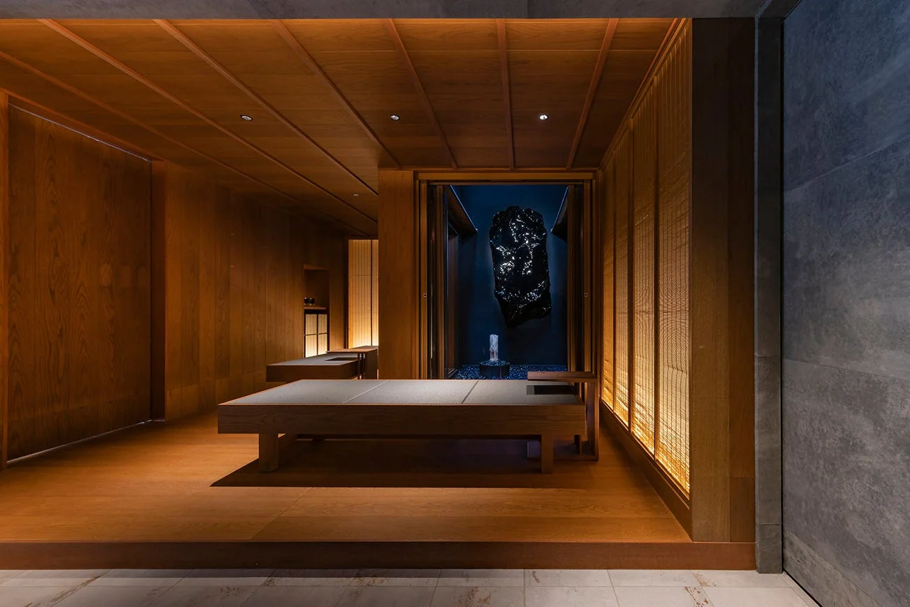 温かい木材で統一されたモダンな和風インテリア空間。低い台が配置され、奥には黒いオブジェと水景が静かに佇む。間接照明が心地よい、禅を感じさせるリラックス空間です。