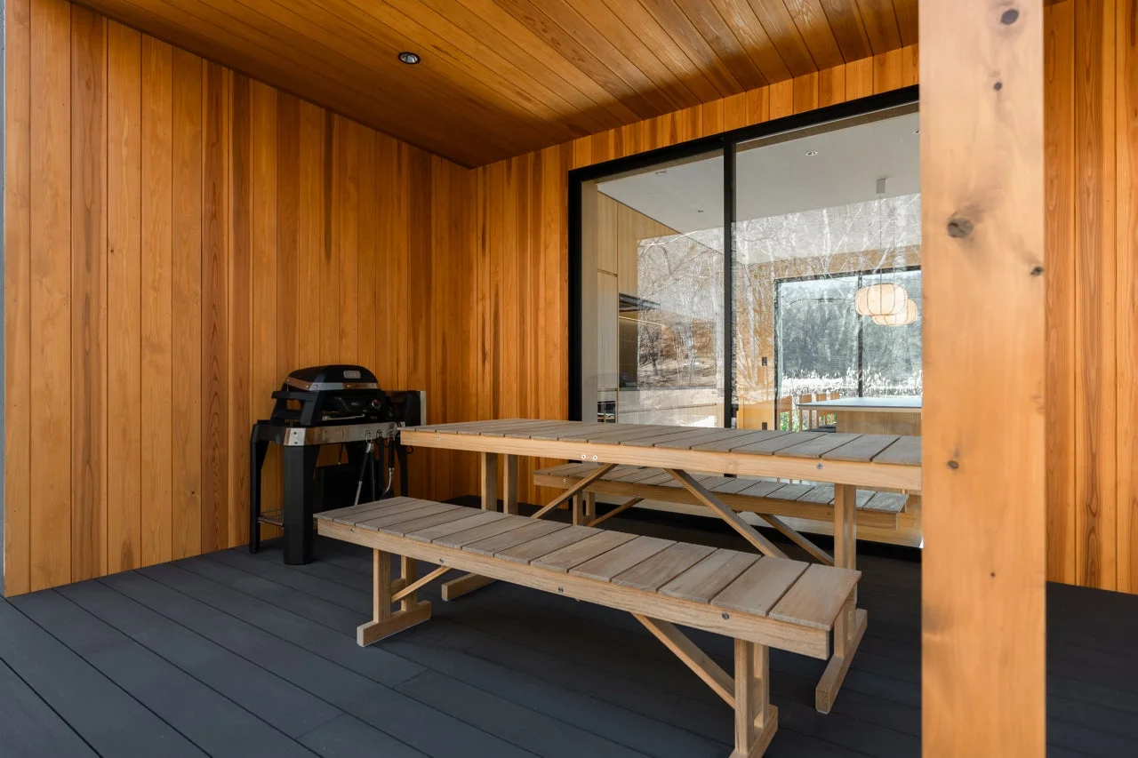 木製の壁と天井が特徴的なモダンなウッドデッキ。バーベキューグリルとピクニックテーブルが設置され、大きな窓からは自然豊かな景色が広がる開放的な空間です。
