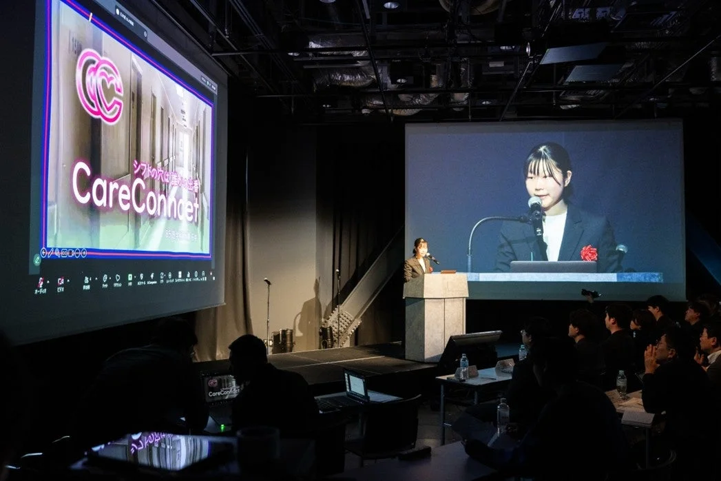 暗い会場で行われているプレゼンテーションの様子。ステージには演台に立つ登壇者がおり、2つの大きなスクリーンが設置されている。左のスクリーンには「CareConfidet」という文字と「シフトの穴は誰が埋めるの？」というテキストが表示され、右のスクリーンには別の登壇者の顔が映し出されている。聴衆が熱心に発表を見守っている。