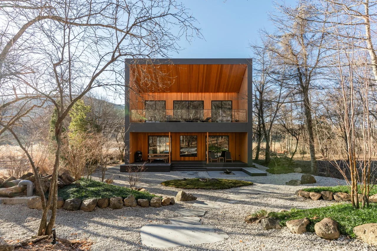 自然豊かな森の中に建つ、木材とダークグレーの外壁が特徴的なモダンな二階建て住宅です。手前には砂利と石、苔で構成された手入れの行き届いた庭が広がり、落ち着いた雰囲気を醸し出しています。