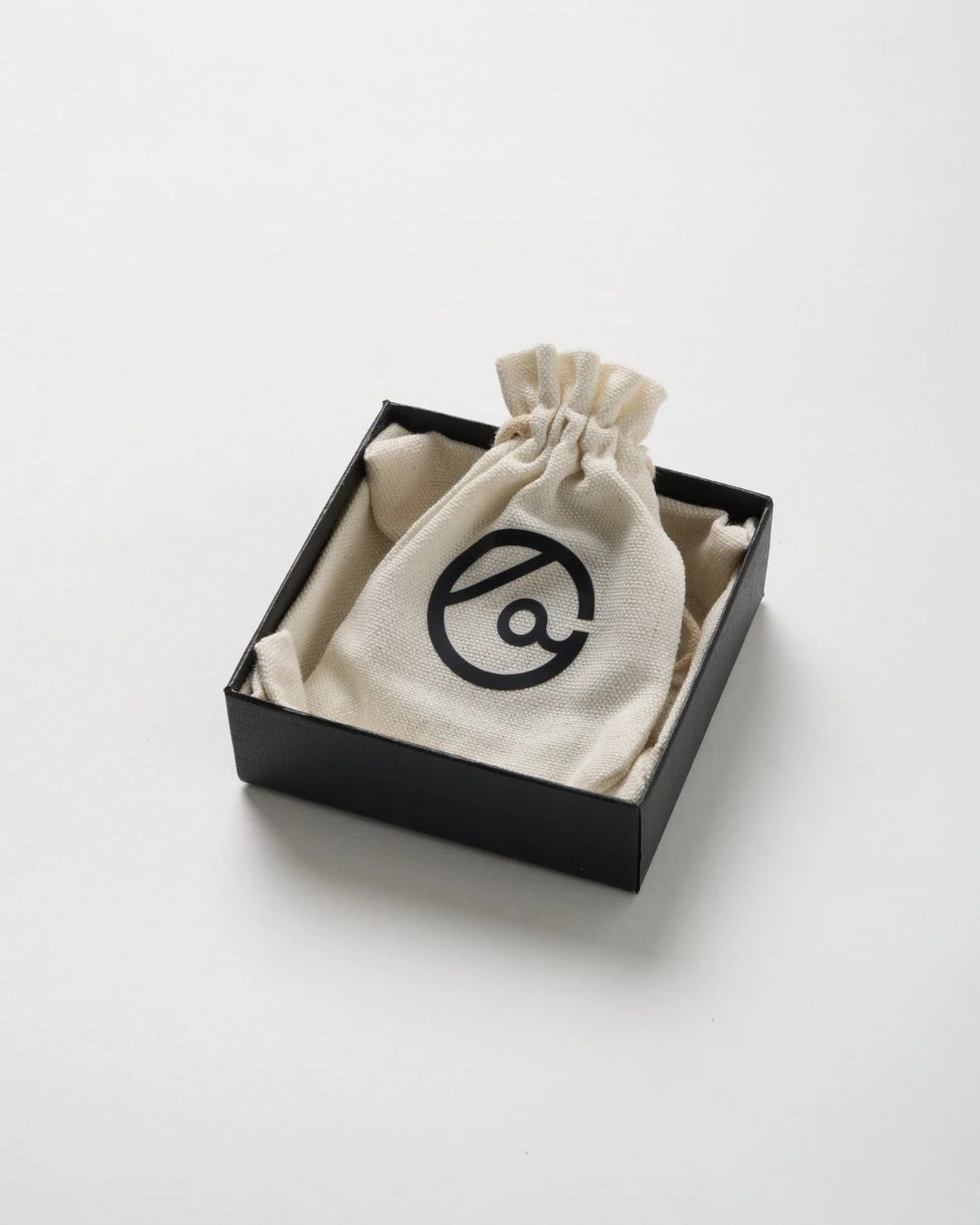 黒い箱の中にベージュの巾着袋が入っており、袋には黒い抽象的なロゴマークが印刷されています