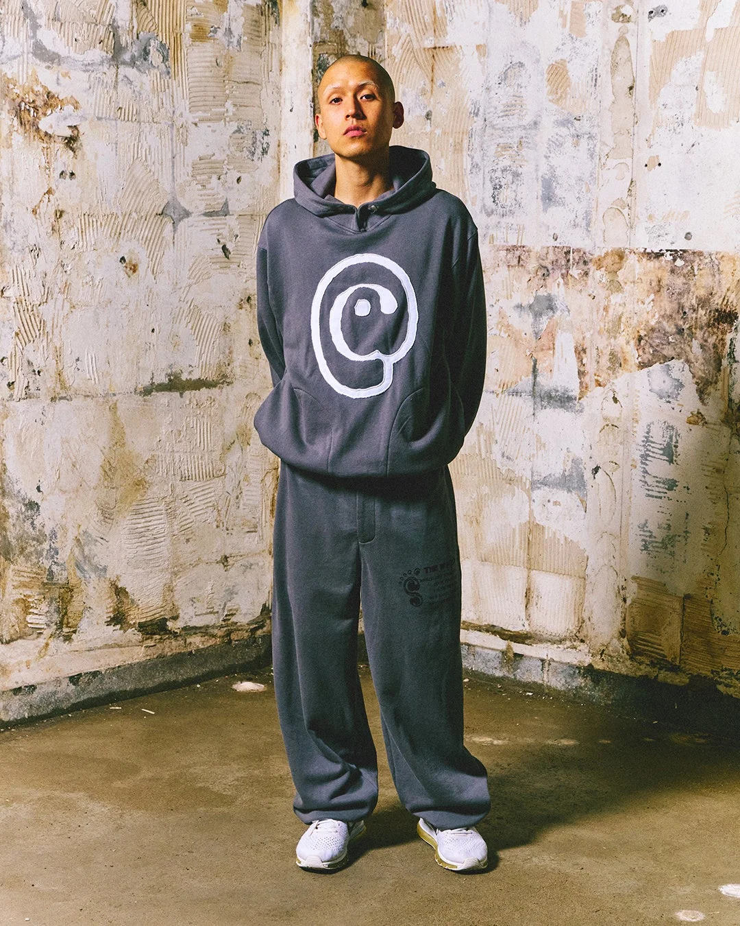 グレーのフーディーとスウェットパンツを着用した男性が、荒廃した壁の前で立っている全身像