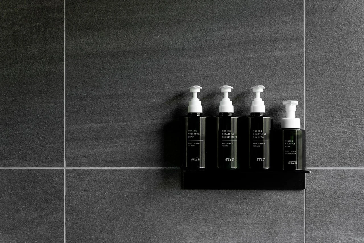 黒いボトルに入ったOSAJIのシャンプー、コンディショナー、ソープ、マルチウォッシュがバスルームの壁に設置されたラックに整然と並べられています。モダンでミニマルなバスアメニティのセットです。