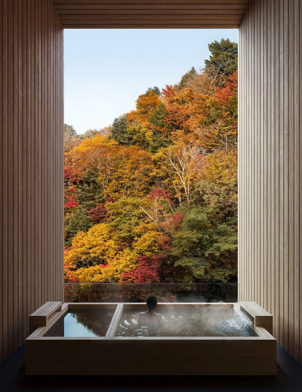 モダンな木製露天風呂で、色鮮やかな紅葉の山々を眺めながら入浴する人物の姿。