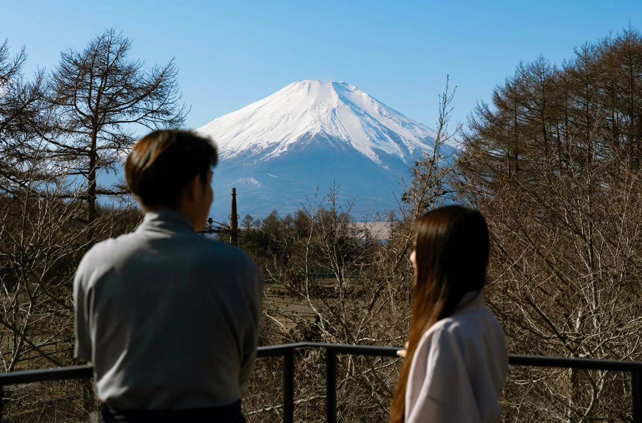 冬の晴れた日、バルコニーから雪化粧した雄大な富士山を眺める二人の後ろ姿。澄み切った青空の下、広がる自然豊かな日本の美しい景色が印象的です。