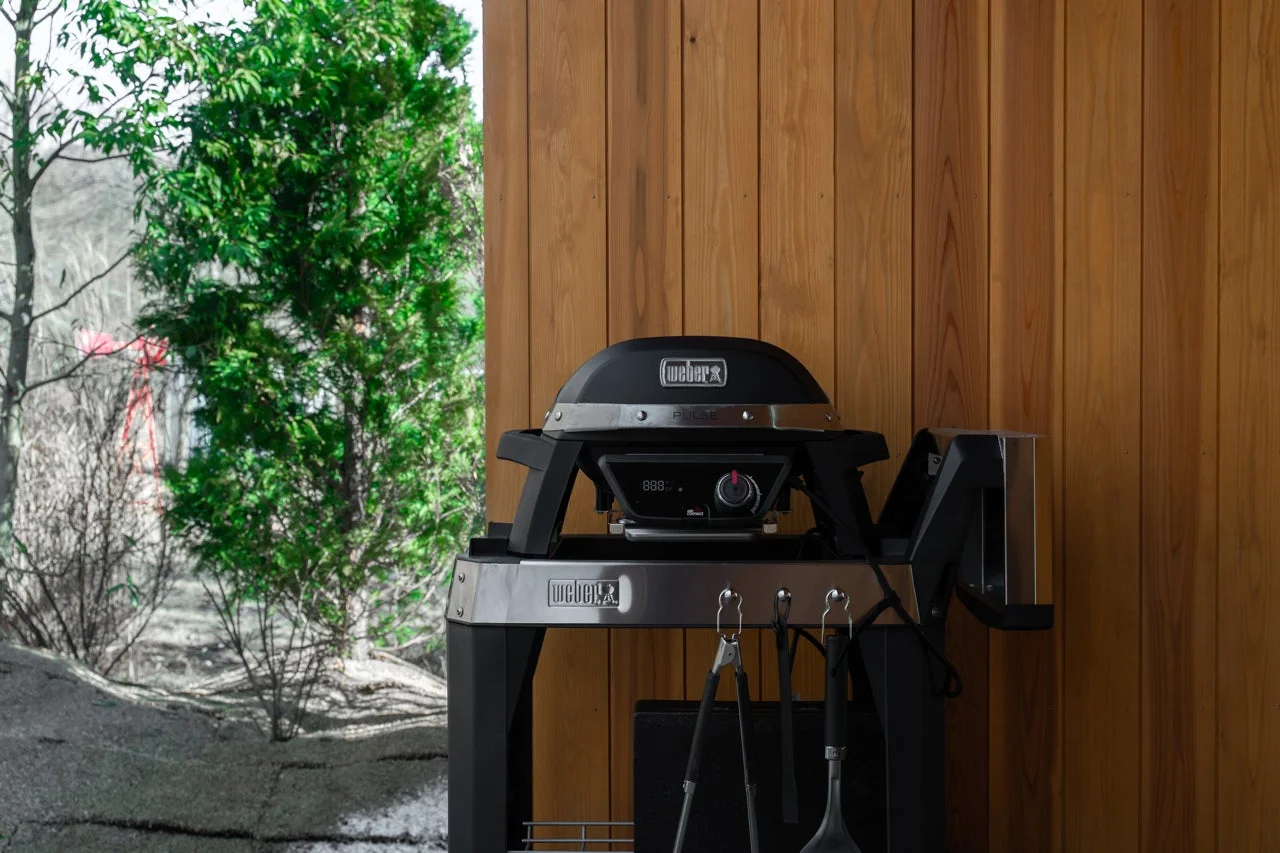 木製の壁を背景に、黒いWeber Pulse電気グリルが屋外に設置されています。グリルには調理器具が吊るされており、背景には緑豊かな木々が見える、アウトドアでのバーベキューシーンを捉えた一枚です。