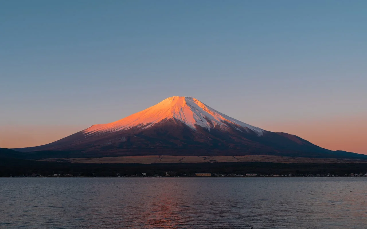 雪を冠した富士山が、夜明けか夕暮れ時の太陽の光を浴びて山頂付近が赤く染まっている雄大な風景です。手前には湖が広がり、その水面に空と山の色が映り込んでいます。