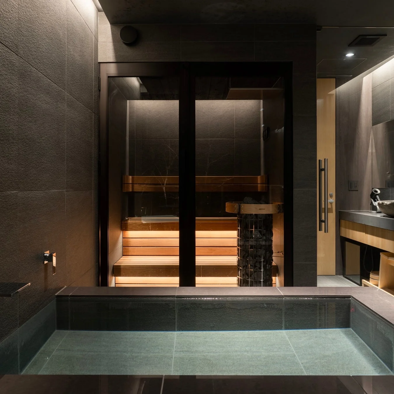 モダンで落ち着いた雰囲気のバスルームとプライベートサウナ。ダークトーンのタイルと温かみのある照明が特徴で、リラックスできる空間を演出している。