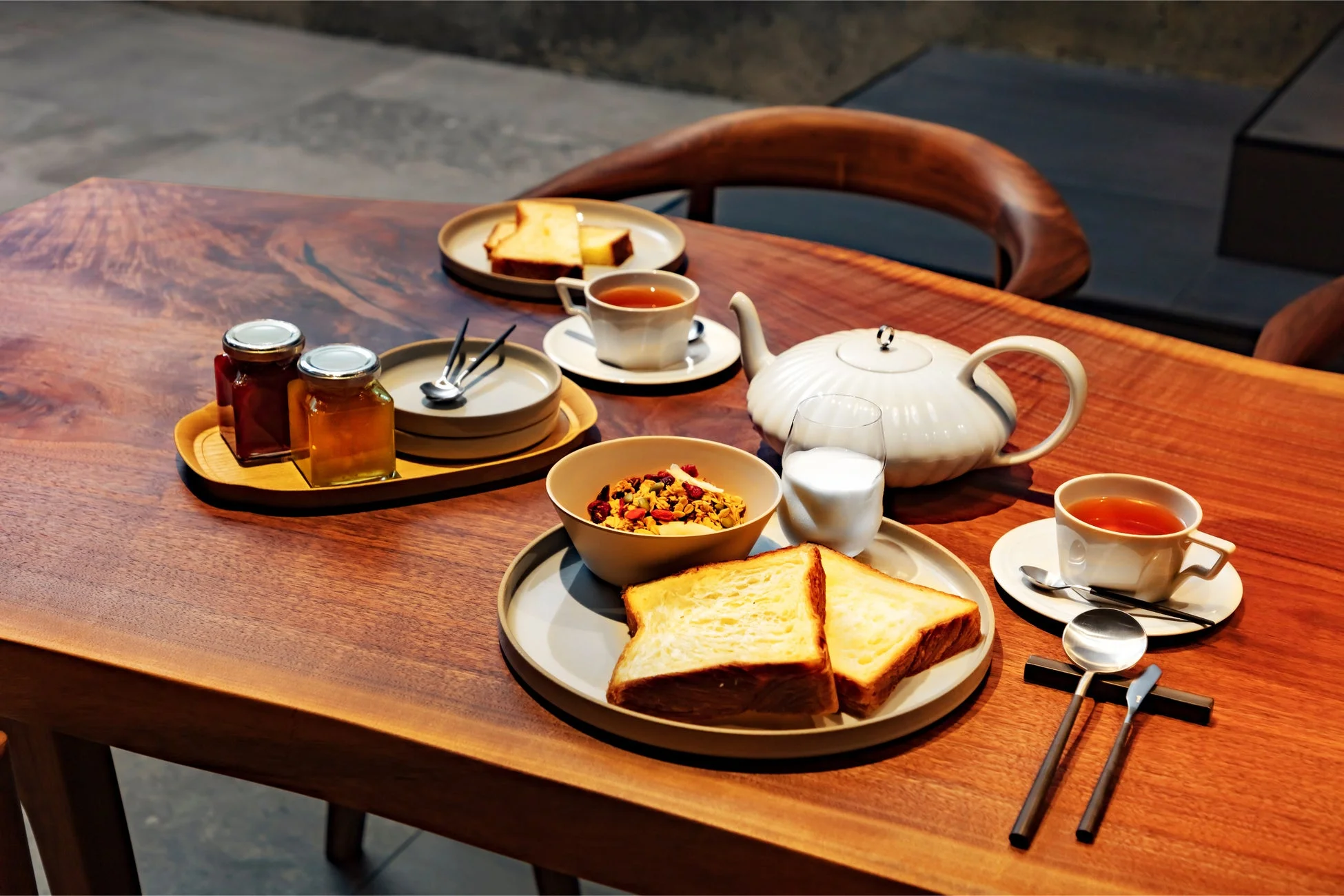 温かい木製のテーブルに、トースト、グラノーラ、紅茶、ミルクなどが並べられた朝食の風景。ジャムや蜂蜜の瓶も添えられ、ゆったりとした食事の時間を演出しています。