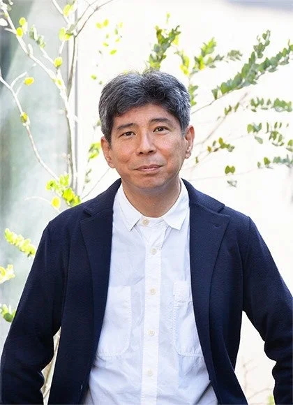 紺色のジャケットと白いシャツを着た中年の日本人男性が、屋外の明るい背景と植物の前で穏やかに微笑んでいるポートレート写真です。