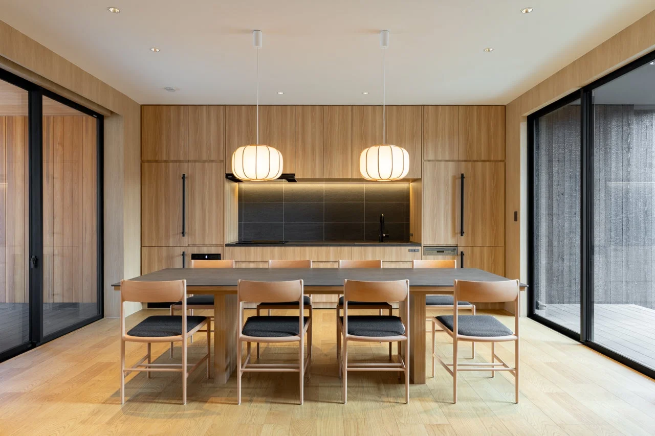 木目調のモダンなダイニングキッチン。中央に長いダイニングテーブルと椅子、上部に特徴的なペンダントライトが2つ。木製キャビネットと黒いキッチンカウンターが調和し、開放的な窓が明るい広々とした空間を演出しています。