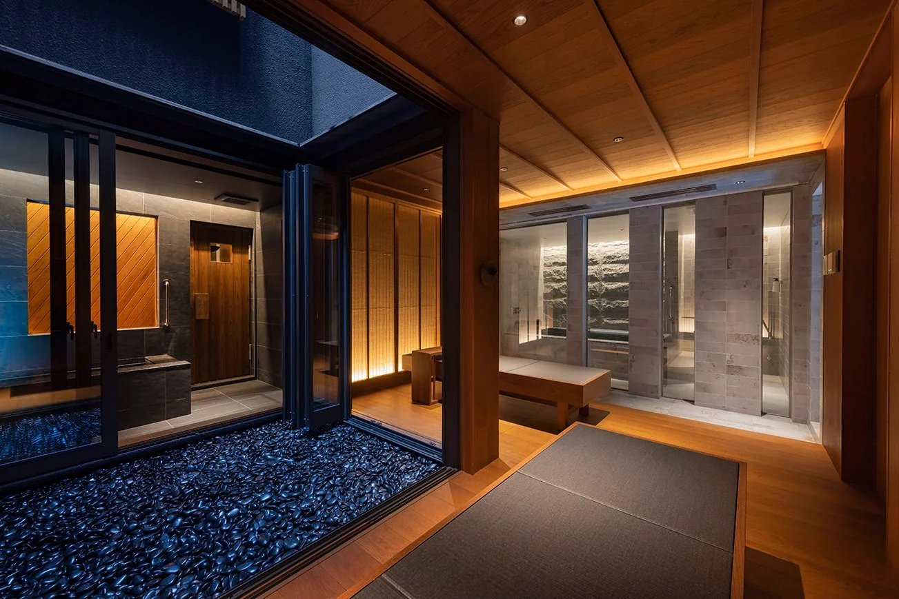 モダンな和風デザインが特徴の高級スパ施設。木材と石を基調とした落ち着いた空間で、間接照明が心地よい雰囲気を演出しています。屋外には黒い小石が敷き詰められた坪庭が見えます。