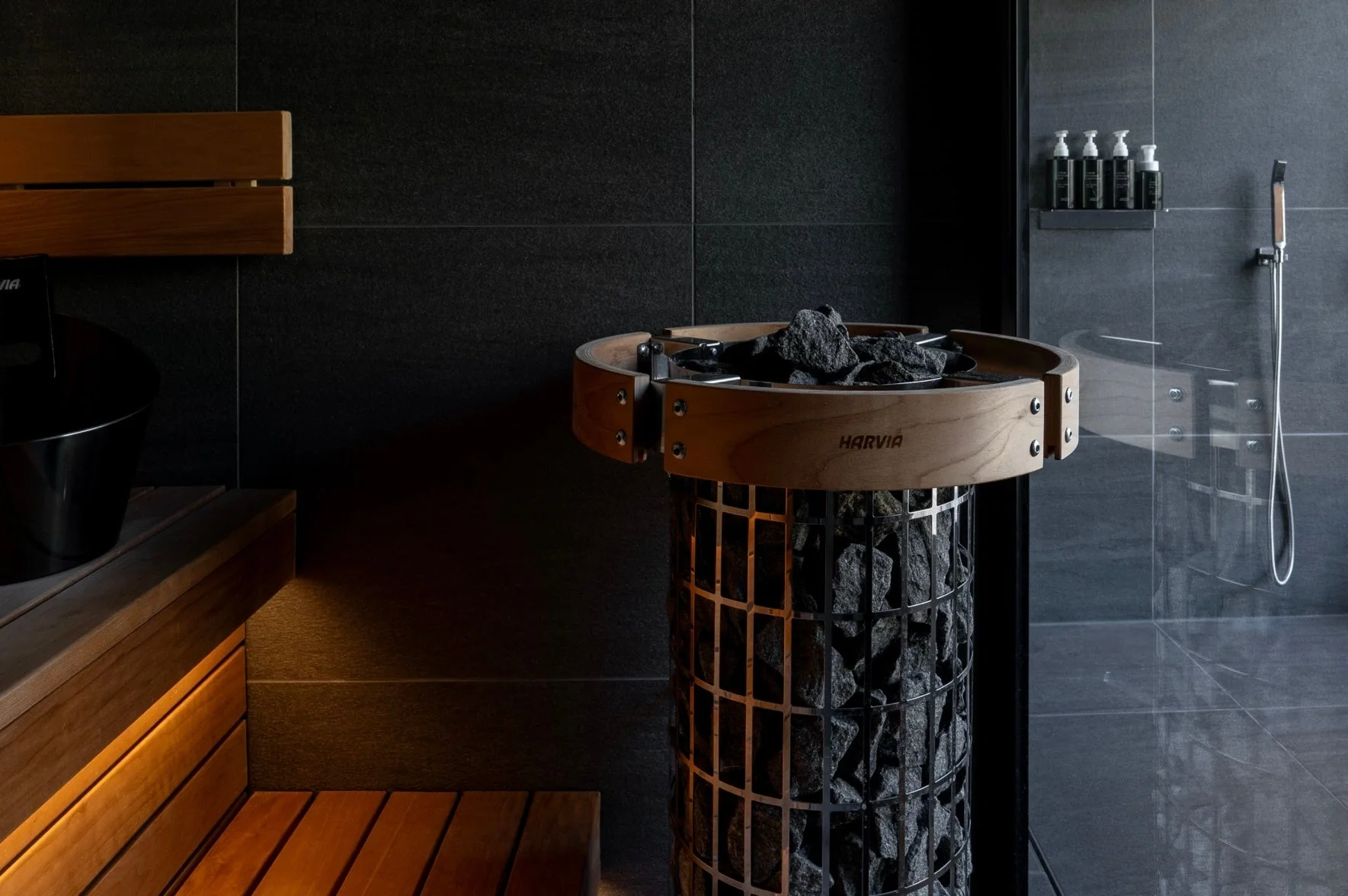 モダンなデザインのサウナ室で、木製のベンチとスタイリッシュなサウナストーブが特徴です。隣にはシャワーエリアも見え、リラックスできる空間が演出されています。