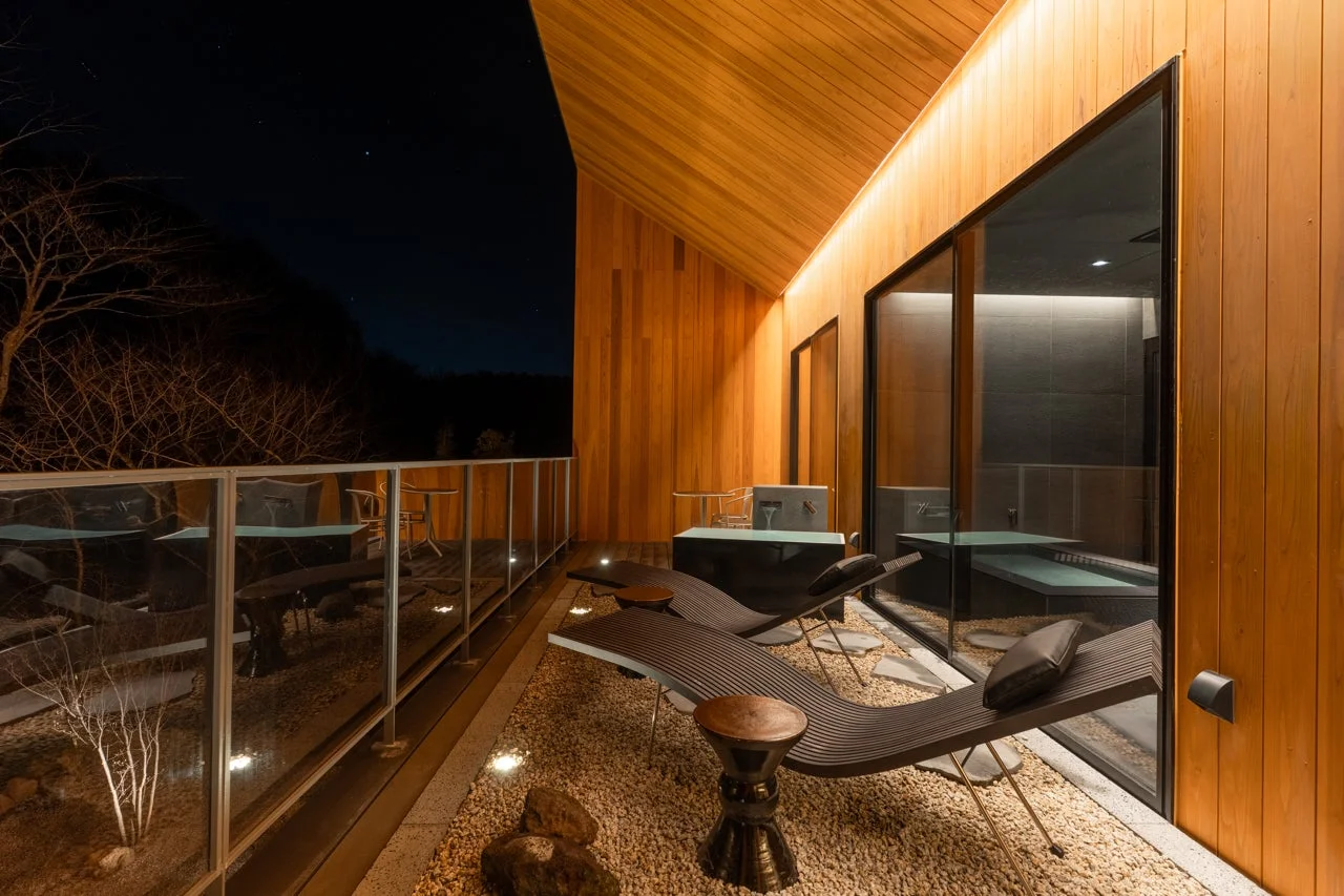 夜のモダンな宿泊施設のテラスで、星空の下、木製の壁とガラスの手すりに囲まれた空間が広がっています。くつろげるラウンジチェアが配され、窓越しにはプライベートな露天風呂が見える、贅沢で落ち着いた雰囲気を醸し出しています。