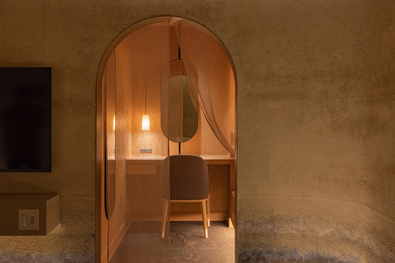 土壁のような質感の壁に囲まれた空間に、アーチ状の入り口がある書斎スペース。デスク、椅子、楕円形の鏡、温かい光を放つペンダントライトが配置され、落ち着いた雰囲気を醸し出すモダンなインテリアです。