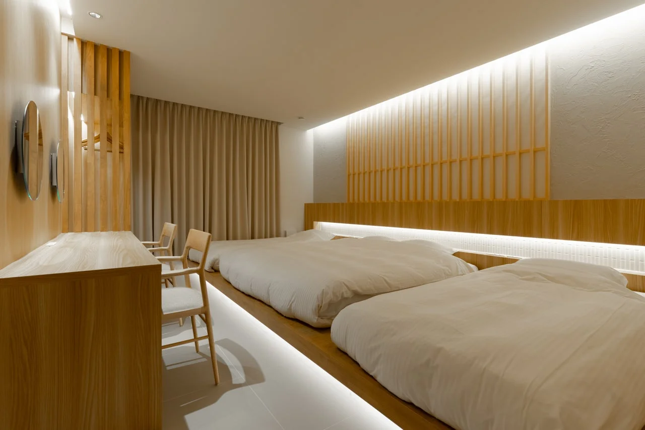 木と白を基調としたモダンでミニマルな寝室の様子です。床上げされたベッドスペースに2つのベッドが並び、間接照明が温かい雰囲気を演出しています。和の要素も感じさせる、落ち着いた快適な空間です。